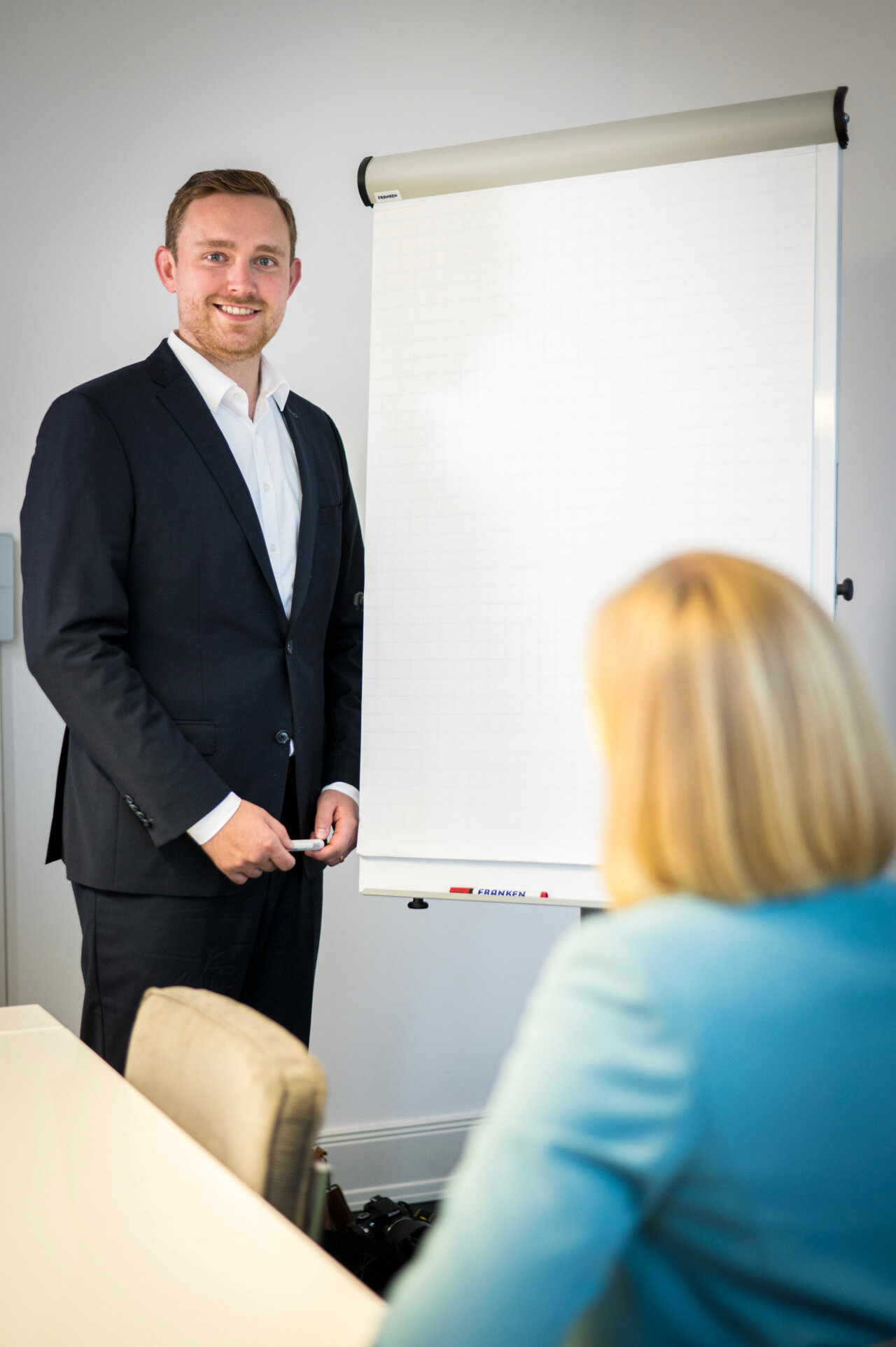 Ein lächelnder EGIDO-Mitarbeiter in einem Anzug steht neben einem leeren Flipchart und hält einen Stift in der Hand. Eine Person mit blonden Haaren ist im Hintergrund sichtbar und scheint ihm zuzuhören.