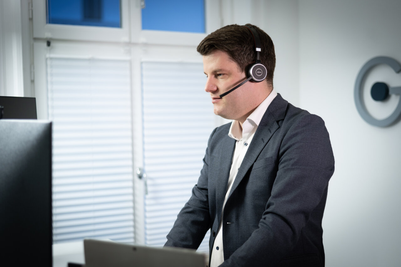 Ein EGIDO-Mitarbeiter in einem dunklen Anzug trägt ein Headset mit Mikrofon und sieht konzentriert aus, während er an einem Schreibtisch mit einem Laptop und einem Monitor arbeitet. Im Hintergrund sind Fenster mit Jalousien sichtbar.