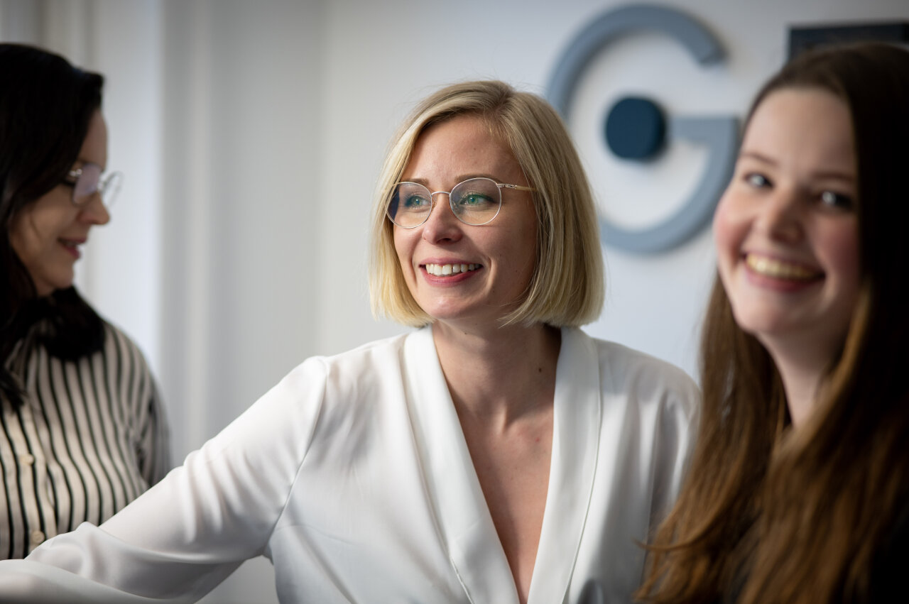 Drei EGIDO-Mitarbeiterinnen stehen in einem hellen, modernen Büro. Eine blonde EGIDO-Mitarbeiterin mit Brille lächelt freundlich in die Kamera, während die anderen beiden Mitarbeiterinnen, eine mit dunklen Haaren und die andere mit langen, braunen Haaren, im Hintergrund leicht verschwommen zu sehen sind und ebenfalls lächeln.