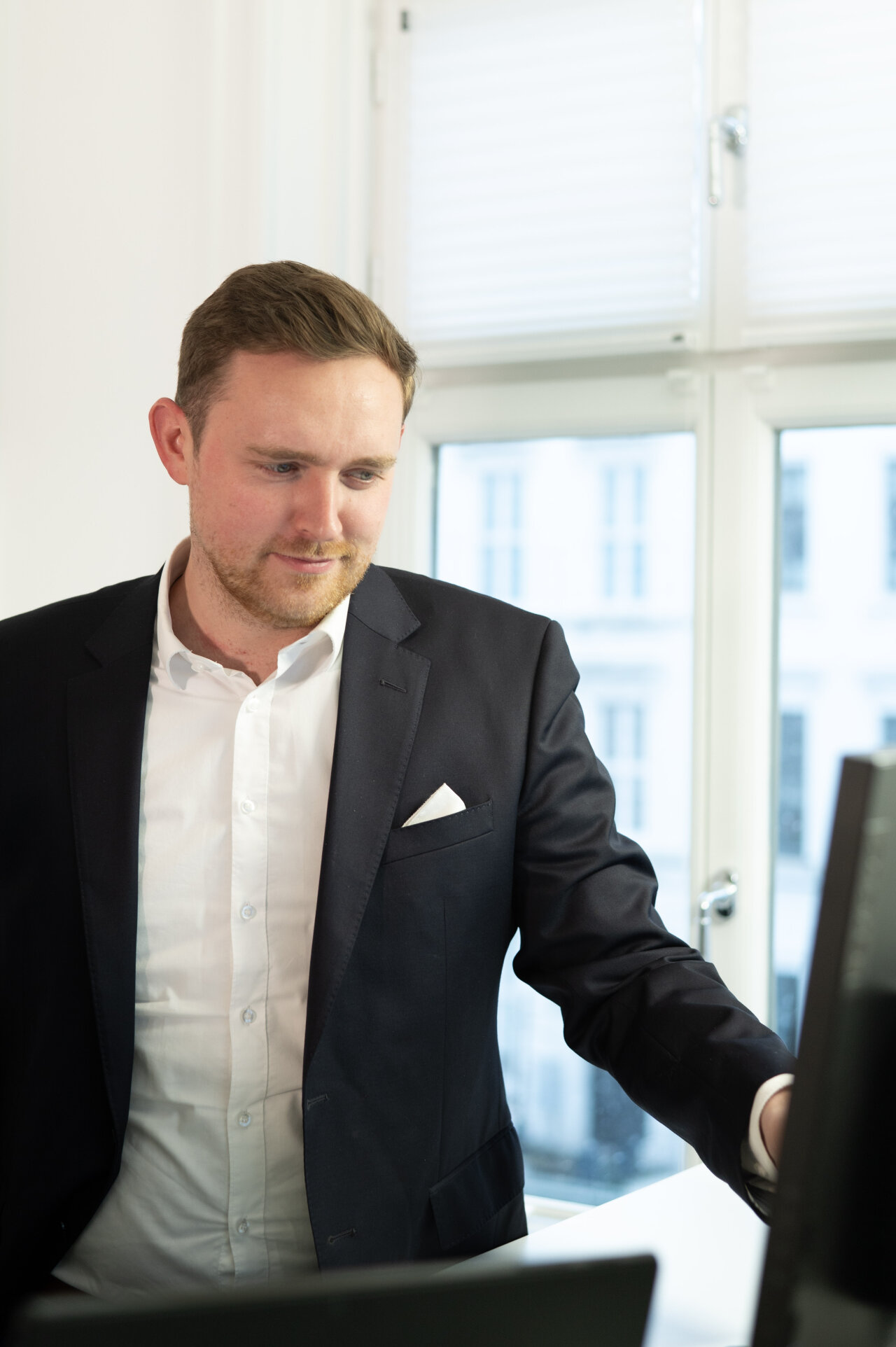 Ein EGIDO-Mitarbeiter in einem dunklen Anzug steht an einem Schreibtisch und schaut auf einen Computerbildschirm. Er hat kurzes, blondes Haar und trägt ein weißes Hemd mit einem dunklen Jacket. Im Hintergrund sind Fenster mit Jalousien zu sehen, die Licht in den Raum lassen.