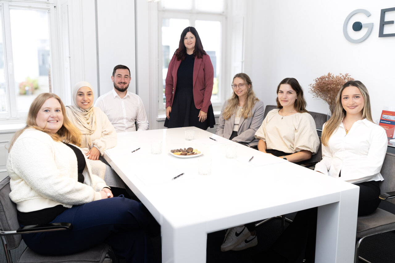 Eine Gruppe von sieben EGIDO-Mitarbeitern sitzt an einem modernen Tisch in einem hellen, eleganten Büroraum. Sechs Personen sind in formeller, business-tauglicher Kleidung gekleidet, während eine Person steht. Auf dem Tisch liegt ein Teller mit Snacks und einige Schreibutensilien. Im Hintergrund sind große Fenster und dekorative Elemente sichtbar.